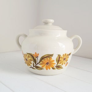 May include: White ceramic crock with a lid, featuring a floral design of yellow and brown flowers on a vine.
