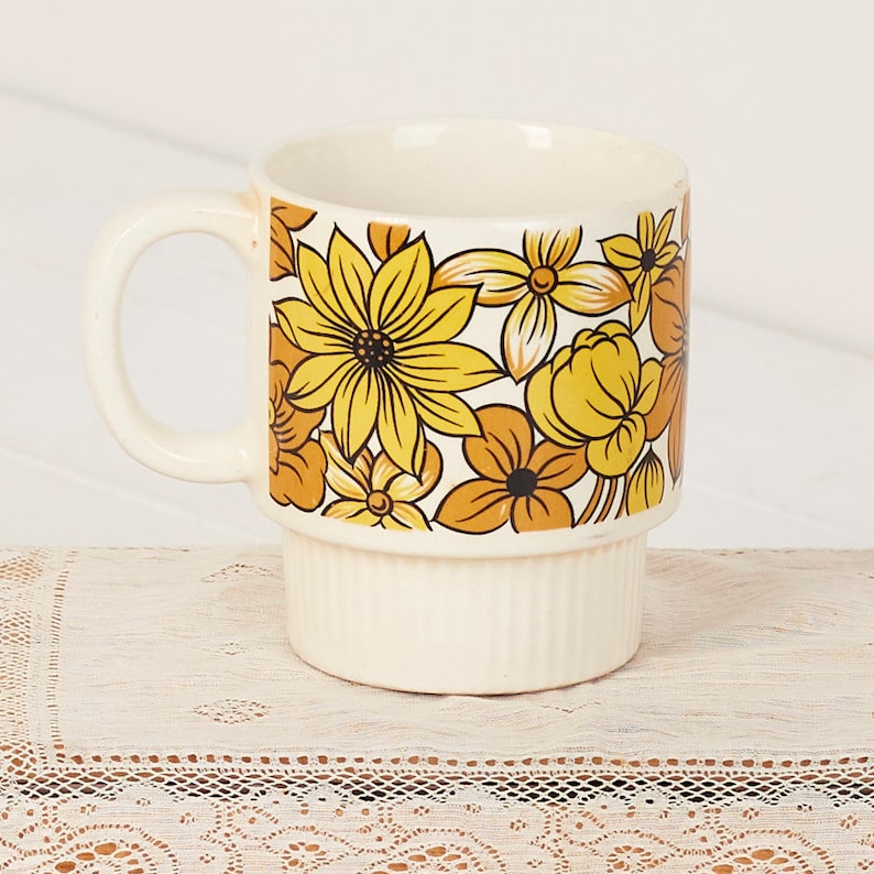Op de afbeelding: Een witte keramische mok met een geel en oranje bloemenpatroon. De mok heeft een brede, afgeronde handgreep en een gestapelde vorm.