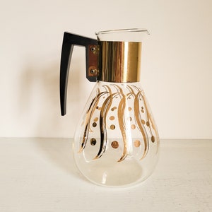 May include: A glass coffee pot with a black handle and gold accents. The pot has a gold and white pattern of stylized leaves and dots.