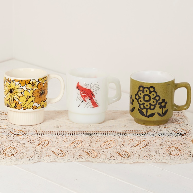 Op de afbeelding: Drie vintage keramische mokken met verschillende bloemenpatronen. De mokken zijn wit met een geel, oranje en bruin bloemenpatroon, een witte mok met een rode kardinaal en een groene mok met een zwart bloemenpatroon.