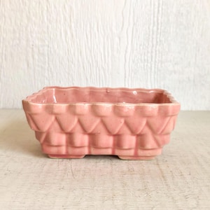 May include: A rectangular, pink ceramic planter with a textured, basketweave-like design. The planter has a scalloped rim and sits on four small feet. The background is a light-colored, textured surface.