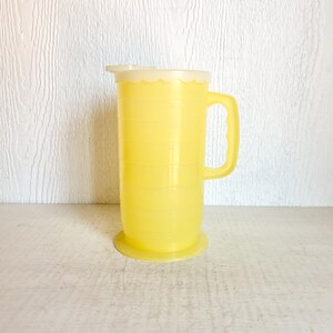 May include: A vintage yellow plastic pitcher with a handle and a white lid. The pitcher has measurement markings and a scalloped edge on the lid. The pitcher is sitting on a light-colored surface.