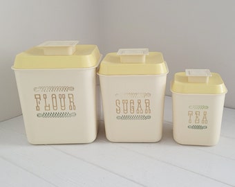 May include: Set of three vintage kitchen canisters with yellow lids and white bodies. The canisters are labeled "Flour", "Sugar", and "Tea" in a decorative font.