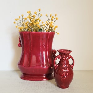 May include: Two red ceramic vases with a floral design. The larger vase has a wide mouth and two handles. The smaller vase has a narrow mouth and two handles. Both vases are filled with yellow flowers.