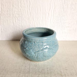 May include: A small, round, light blue ceramic pot with a textured surface. The pot has a wide opening and a slightly uneven rim. The exterior features a raised, organic pattern. This decorative pot is suitable for small plants or as a decorative item.