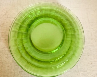May include: A round, transparent, lime green glass plate. The plate has a central recessed area and concentric ridged rings around the edge. The glass has a slight sheen, reflecting light. The plate is likely for serving food.