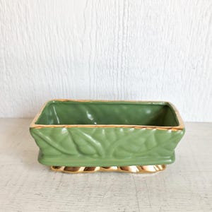 May include: A rectangular, green ceramic planter with gold trim. The planter features a leaf design on the sides and a gold base. The interior is a solid green color. The planter is suitable for small plants.
