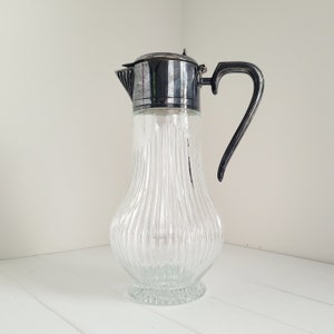 May include: A clear glass pitcher with a silver lid and handle. The pitcher has a ribbed design and a round base.