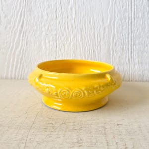 May include: A small, yellow ceramic bowl with a decorative floral design. The bowl has a rounded base and a slightly flared rim.