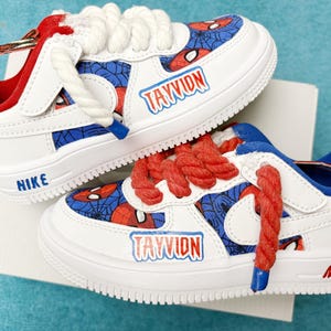 May include: A pair of white sneakers with red and blue Spiderman themed designs. The sneakers have the name "TAYVION" written on them and the Nike logo on the side.