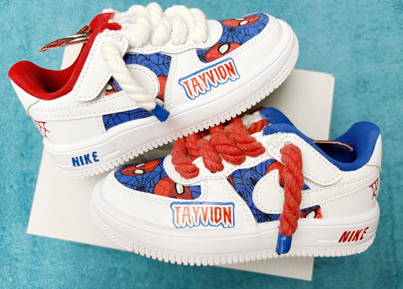 May include: A pair of white sneakers with red and blue Spiderman themed designs. The sneakers have the name "TAYVION" written on them and the Nike logo on the side.