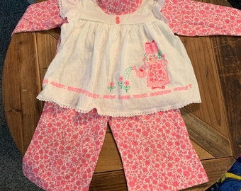Puede incluir: Un conjunto de dos piezas blanco y rosa para un bebé o un niño pequeño. La parte superior es un vestido blanco con un cuello de estampado floral rosa y mangas largas. El vestido tiene una imagen bordada rosa de una niña regando flores con el texto "Oh Mary, Country Girl, How Does Your Garden Grow?" La parte inferior es un pantalón rosa con un estampado floral blanco.