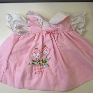 Puede incluir: Un vestido rosa sin mangas con cuello blanco y mangas abullonadas. El vestido tiene un diseño de bordado floral con dos flores rosas y hojas verdes.