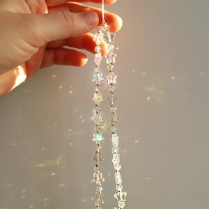 May include: A strand of iridescent star-shaped crystal beads hangs from a white string. The beads are strung on a silver wire and catch the light, creating a rainbow effect.