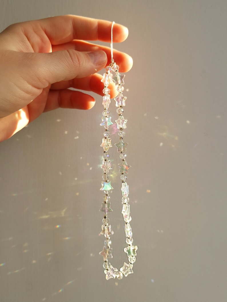 May include: A strand of iridescent star-shaped crystal beads hangs from a white string. The beads are strung on a silver wire and catch the light, creating a rainbow effect.