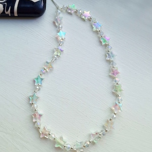 May include: A white phone charm with iridescent star-shaped beads. The charm is attached to a phone case with a thin string.