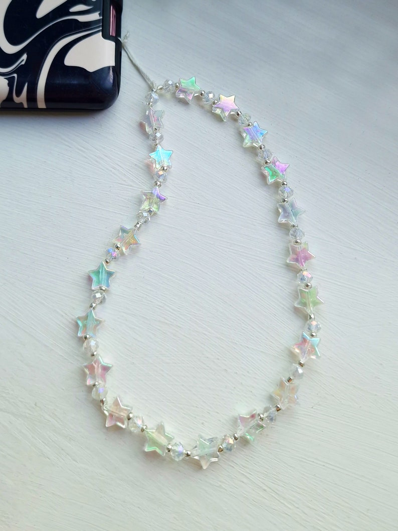 May include: A white phone charm with iridescent star-shaped beads. The charm is attached to a phone case with a thin string.