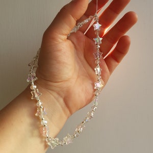 May include: A delicate silver chain necklace with iridescent star-shaped beads. The necklace is held in a hand, showcasing the sparkle of the beads.