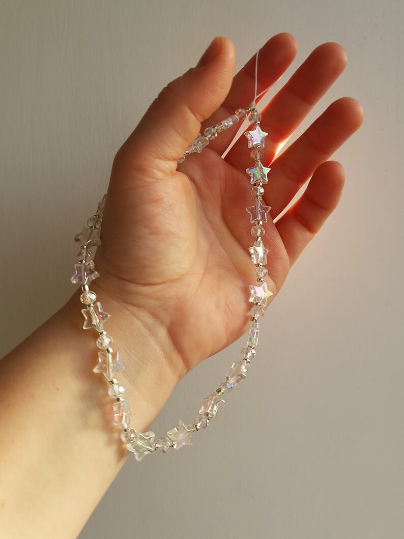 May include: A delicate silver chain necklace with iridescent star-shaped beads. The necklace is held in a hand, showcasing the sparkle of the beads.