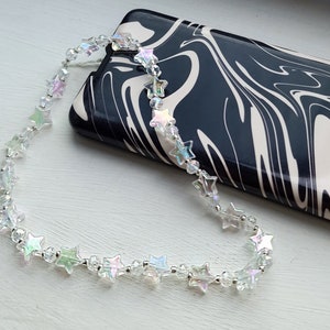 May include: A clear iridescent star-shaped beaded phone charm with a silver chain. The charm is hanging from a black and white marbled phone case.
