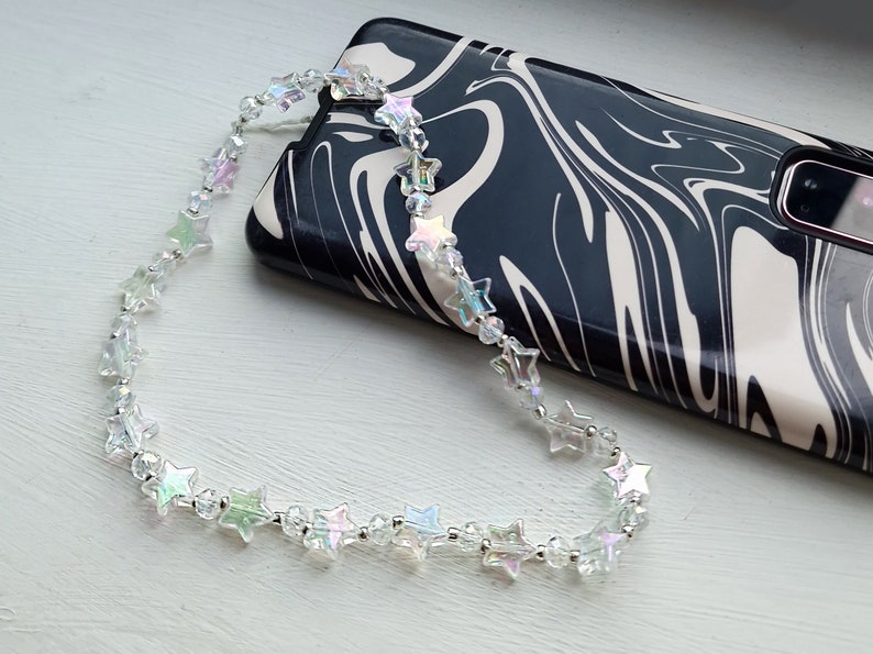 May include: A clear iridescent star-shaped beaded phone charm with a silver chain. The charm is hanging from a black and white marbled phone case.