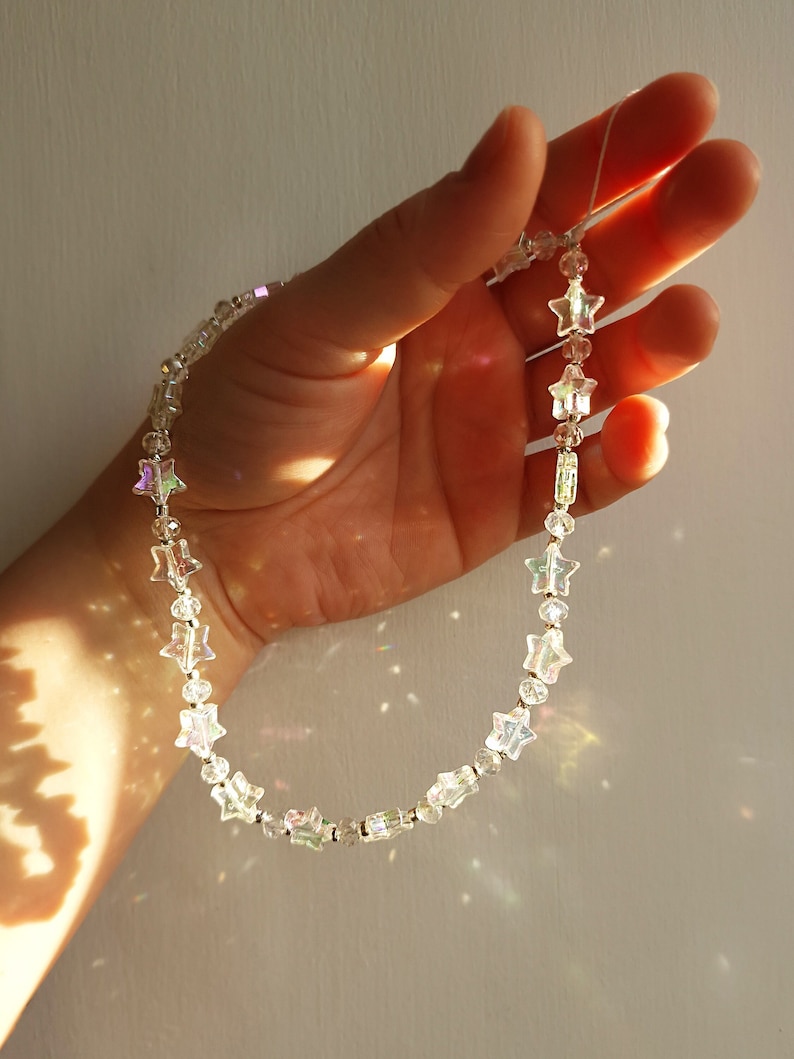 May include: A silver star-shaped beaded necklace with iridescent accents. The necklace is held in a hand against a white background.