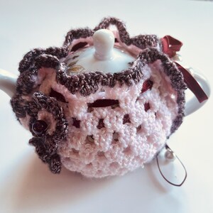 May include: A white teapot with a pink and brown crocheted tea cozy. The cozy has a scalloped edge and a delicate floral pattern.
