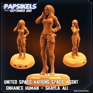 May include: A 3D printed miniature figurine of a female space agent wearing a futuristic outfit and holding a communication device. The figurine is standing on a round base. The text "UNITED SPACE NATIONS SPACE AGENT ENHANCE HUMAN - SHAYLA ALI" is printed below the figurine.