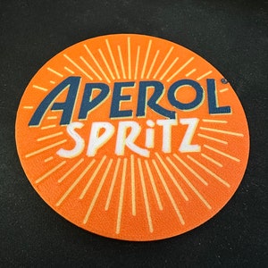 Può includere: Sottobicchiere rotondo arancione con un design a raggiera bianco e il testo "Aperol Spritz" in blu.