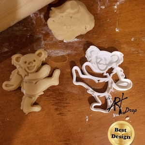 May include: White plastic cookie cutter shaped like a cartoon bear with one leg raised. The bear is smiling and has a flower on its head. The cookie cutter is on a wooden surface with a rolled out piece of cookie dough and a gold award sticker that says "Best Design" with a red ribbon.
