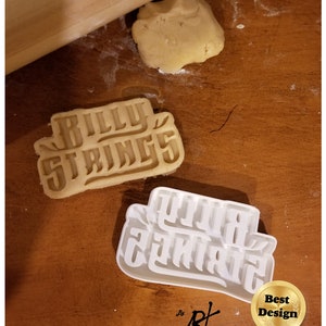 May include: White plastic cookie cutter with the words "Billy Strings" in a stylized font. The cutter is on a wooden surface with a rolled out piece of dough and a rolling pin.
