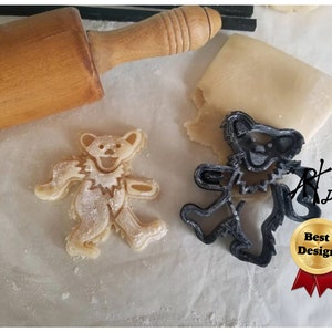 May include: A black plastic cookie cutter in the shape of a dancing bear with a smiling face. The cookie cutter is on a white surface next to a cookie that has been cut out using the cookie cutter.