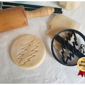 May include: A round cookie cutter with a black lightning bolt design next to a cut-out cookie. A wooden rolling pin and pieces of dough are also visible. The image also includes a "Best Design" badge.