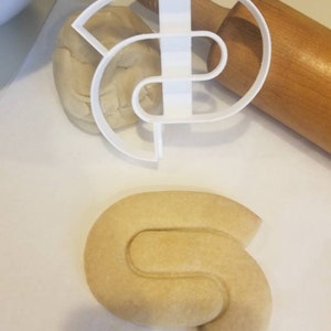 May include: A white plastic cookie cutter shaped like a swirl, with a piece of dough pressed into the cutter. A wooden rolling pin is in the background. A cookie shaped like a swirl is on a white surface.