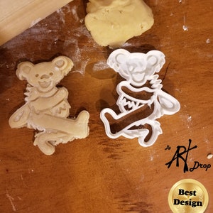 May include: White plastic cookie cutter in the shape of a cartoon bear playing a guitar. The cookie cutter is on a wooden surface next to a brown cookie in the same shape. The cookie cutter is labeled "Art Drop" and "Best Design".