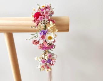 May include: A floral crown featuring a variety of dried flowers in shades of pink, red, purple, and white. The flowers are arranged on a natural-colored base, with a ribbon hanging down. The crown is displayed on a wooden stand.