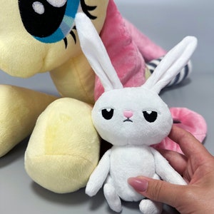 May include: A white plush bunny with a pink nose and black eyes sits next to a yellow and pink plush pony. The bunny has a sad expression.
