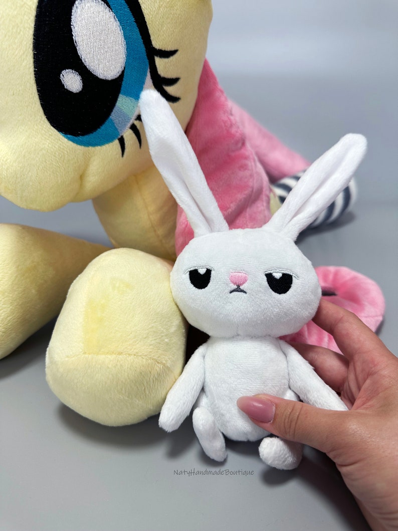 May include: A white plush bunny with a pink nose and black eyes sits next to a yellow and pink plush pony. The bunny has a sad expression.