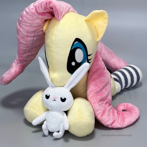 May include: A plush toy of Pinkie Pie from My Little Pony, in her signature yellow and pink colors, with a white plush bunny in her arms. The bunny has a grumpy expression.