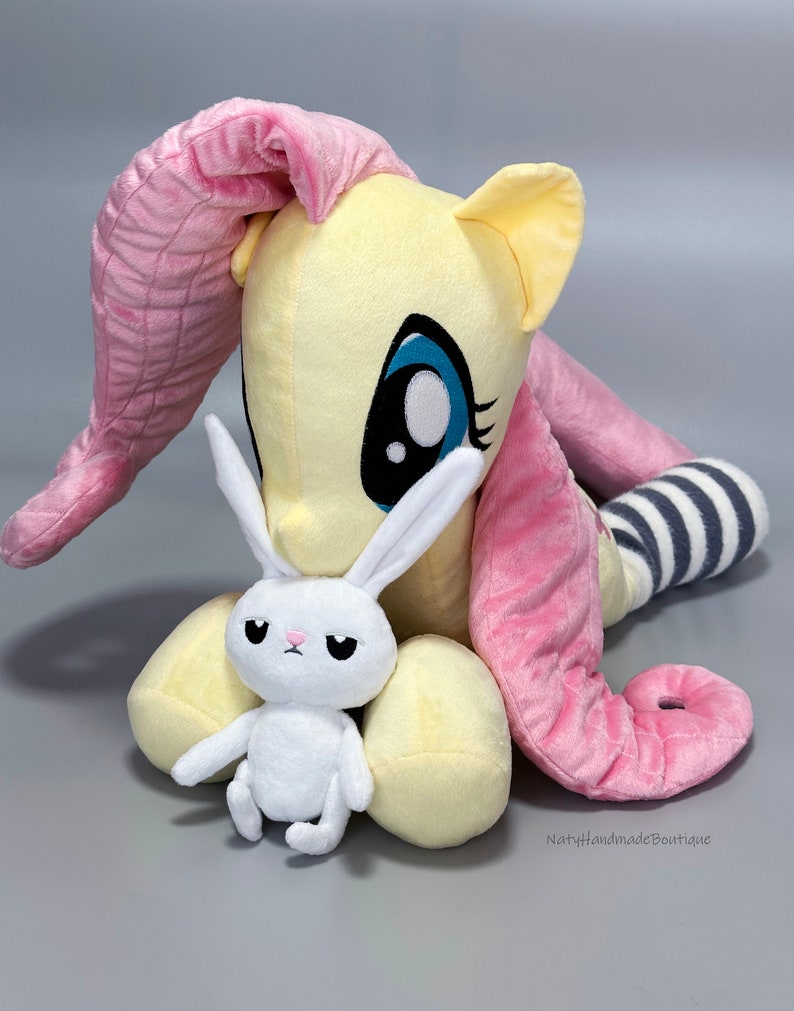 May include: A plush toy of Pinkie Pie from My Little Pony, in her signature yellow and pink colors, with a white plush bunny in her arms. The bunny has a grumpy expression.