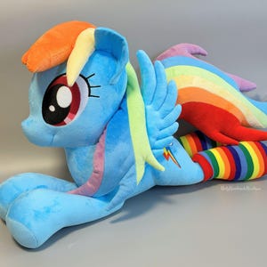 Puede incluir: Un peluche de poni azul con crin y cola de arcoíris multicolor, grandes ojos bordados y alas pequeñas. El poni tiene un símbolo de rayo en el costado y lleva calentadores de piernas a rayas con los colores del arcoíris en las patas traseras.