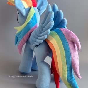 May include: A plush toy of Rainbow Dash from My Little Pony. The toy is blue with a rainbow mane and tail. The toy is standing on a gray surface.