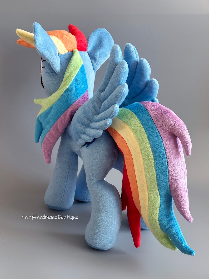 May include: A plush toy of Rainbow Dash from My Little Pony. The toy is blue with a rainbow mane and tail. The toy is standing on a gray surface.