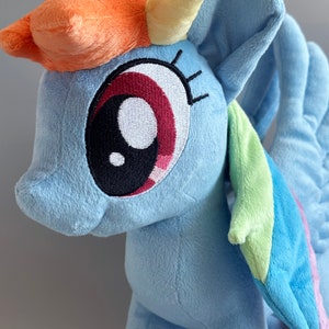May include: A plush toy of Rainbow Dash from My Little Pony. The toy is light blue with a yellow horn, orange and red mane, and a rainbow-colored wing. The toy has a large, embroidered eye with a black pupil and a red iris.