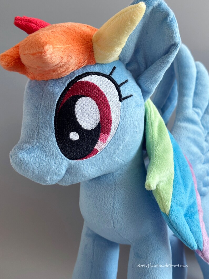 May include: A plush toy of Rainbow Dash from My Little Pony. The toy is light blue with a yellow horn, orange and red mane, and a rainbow-colored wing. The toy has a large, embroidered eye with a black pupil and a red iris.