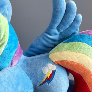 May include: A blue plush toy with a rainbow tail and wings. The toy has a rainbow and cloud design on its side.