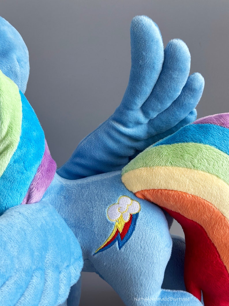 May include: A blue plush toy with a rainbow tail and wings. The toy has a rainbow and cloud design on its side.