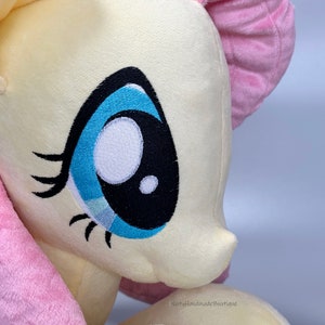 May include: Close-up of a plush toy pony with a yellow body, pink mane and tail, and a blue eye with black eyelashes. The toy is made of soft, fluffy fabric.