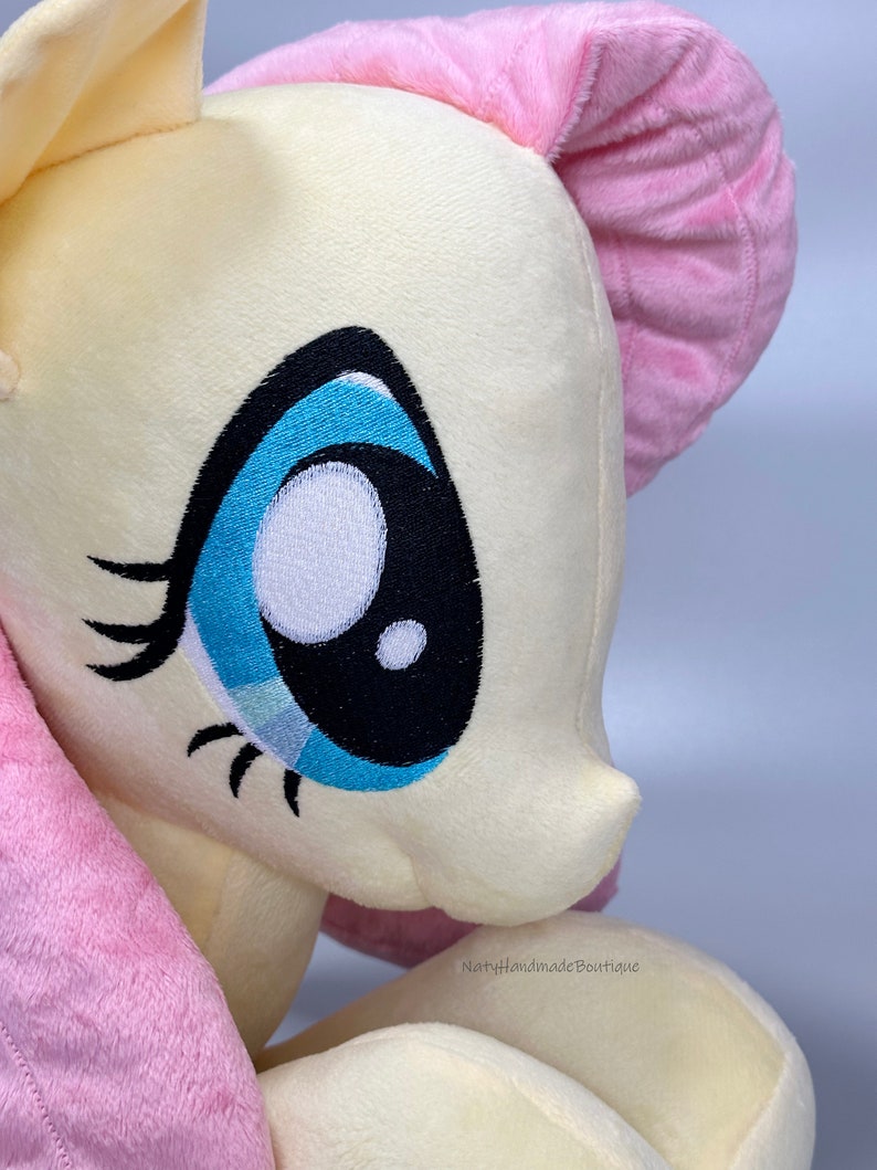 May include: Close-up of a plush toy pony with a yellow body, pink mane and tail, and a blue eye with black eyelashes. The toy is made of soft, fluffy fabric.