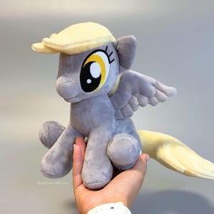 May include: A plush toy of a grey pony with a light yellow mane and tail. The pony features large yellow eyes with black pupils, a pair of small grey wings on its back, and a soft, textured fabric finish. The design is inspired by a cartoon character.